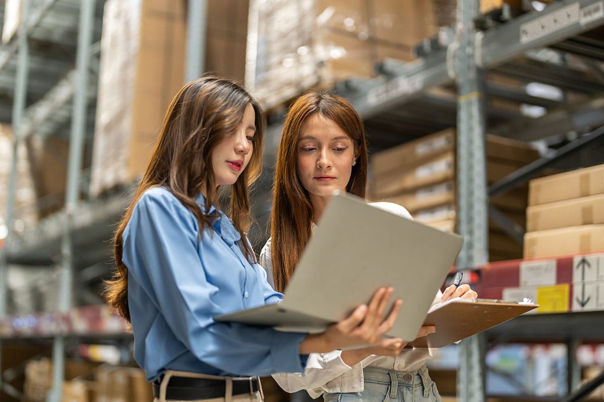 Deux spécialistes de la logistique utilisant un ordinateur portable pour assurer le suivi des stocks à l'intérieur d'un entrepôt doté de hauts rayonnages et de boîtes en carton.