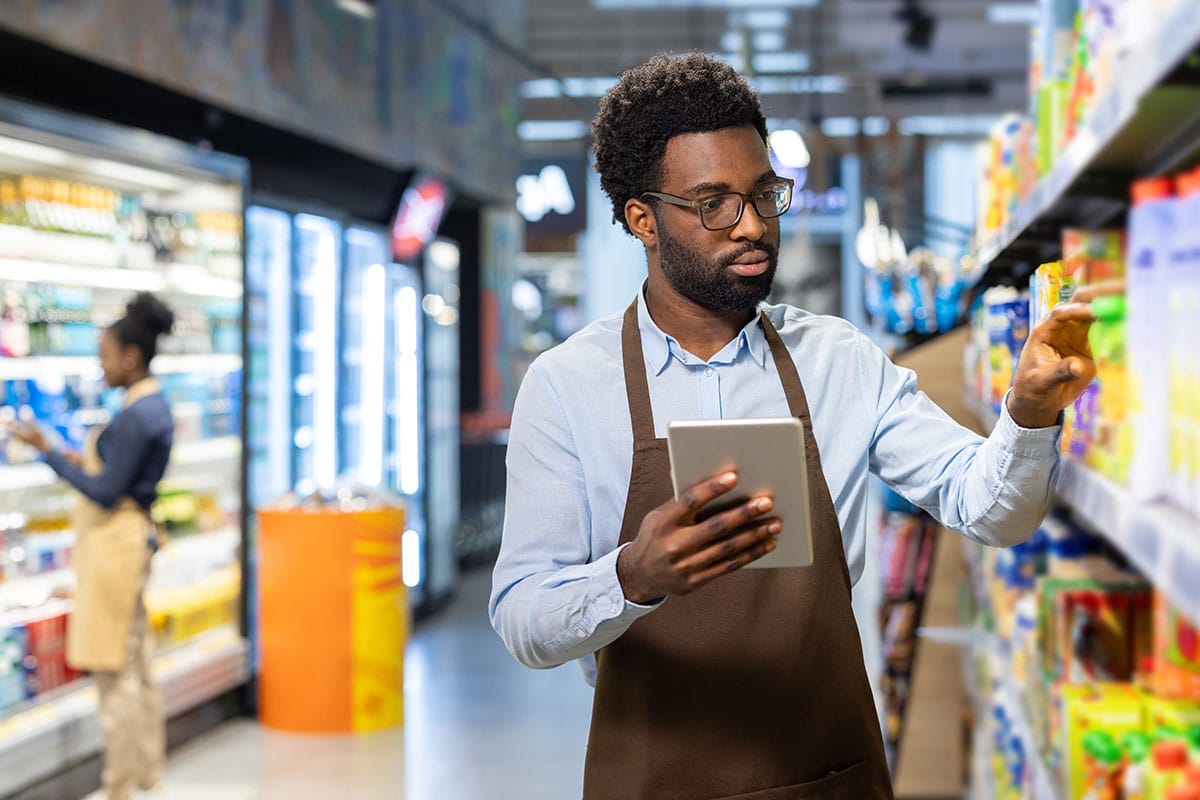 Un employé d’un commerce de détail portant un tablier et tenant une tablette tout en gérant les stocks et en vérifiant les produits sur les rayons d'une épicerie.