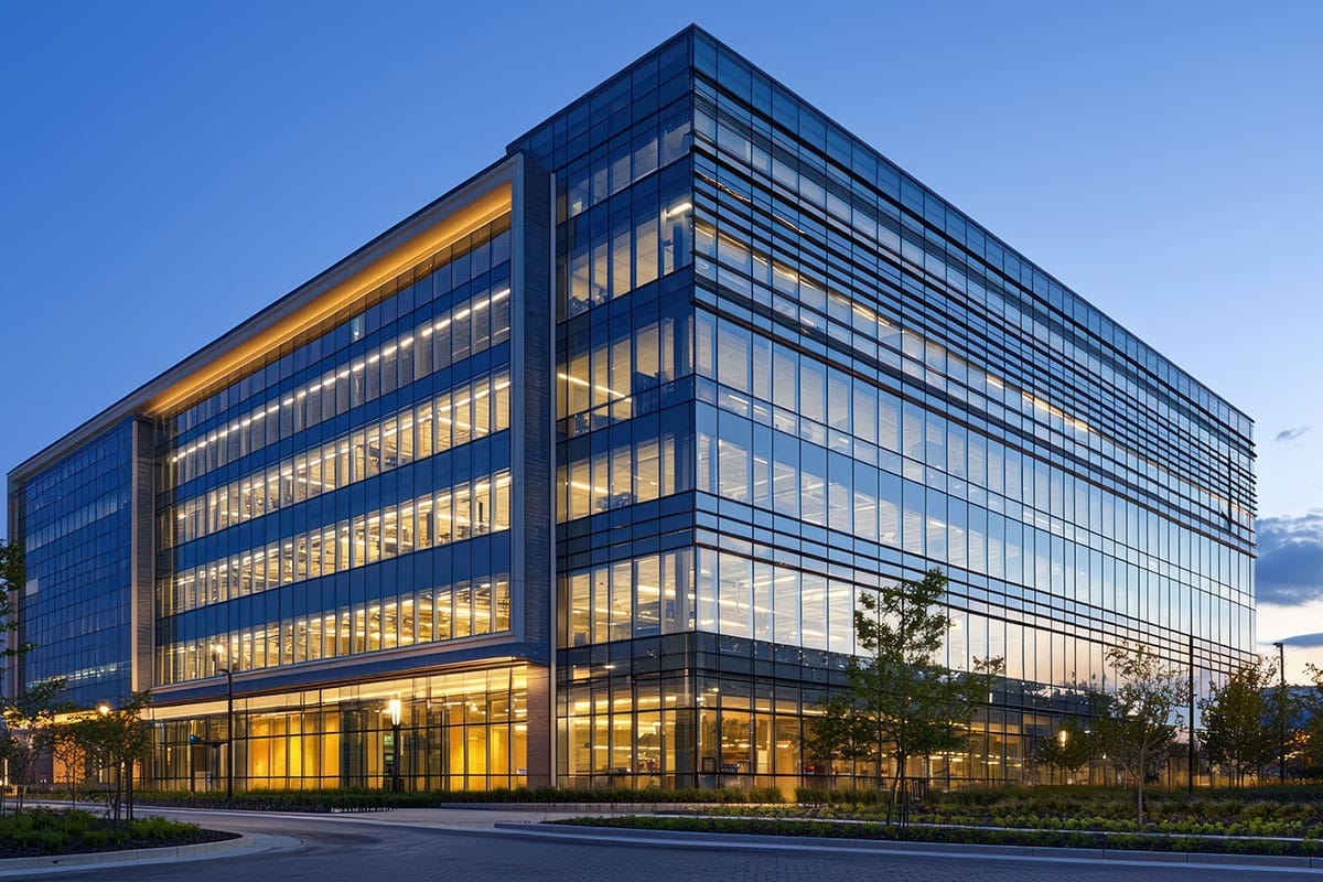 Un immeuble de bureaux moderne en verre à plusieurs étages, illuminé par des lumières intérieures chaleureuses au crépuscule.
