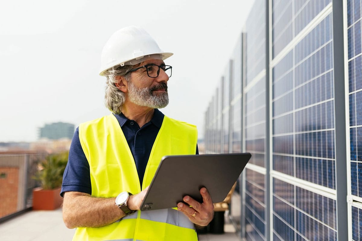 Un travailleur des services publics portant un casque de sécurité blanc et un gilet de sécurité jaune utilise une tablette devant un site de panneaux solaires.
