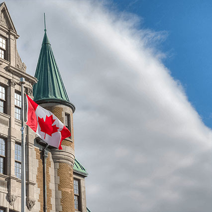 Un drapeau canadien bat au vent à côté d’un immeuble historique du gouvernement fédéral, représentant ainsi des services numériques sécurisés et modernes et des réseaux de confiance soutenant les activités fédérales partout au Canada.