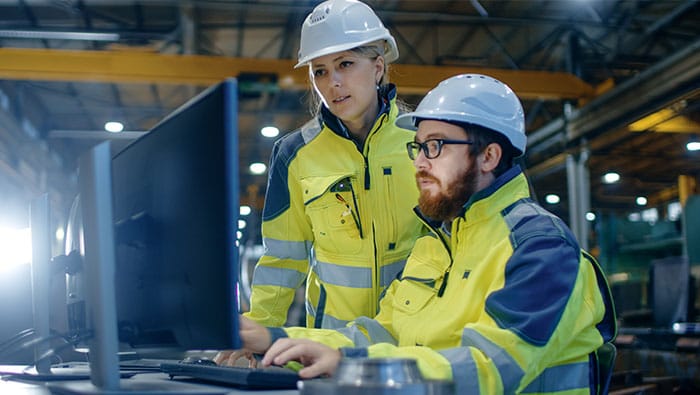 Deux travailleurs dans une usine, portant des casques de sécurité et des gilets de sécurité jaunes, collaborent tout en regardant un ordinateur portable.