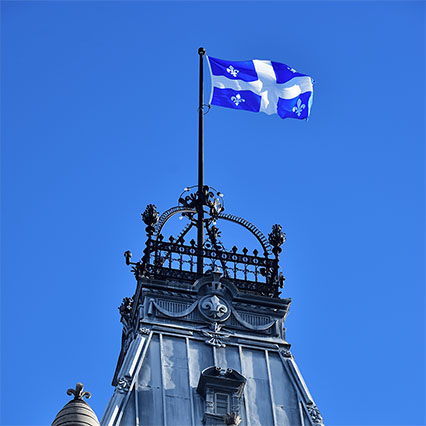 Un drapeau du Québec au sommet d’un édifice du parlement provincial contre un ciel bleu clair, symbolisant une connectivité évolutive et sécurisée et des solutions numériques qui soutiennent les services provinciaux essentiels.